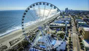 A large Ferris wheel dominates the foreground with a view of a coastal cityscape and beach in bright daylight.