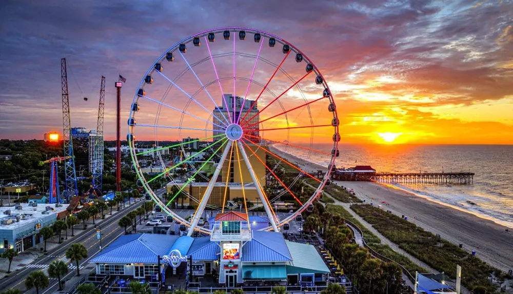 A vibrant sunset paints the sky behind a towering Ferris wheel overlooking a pier along a beachfront adorned with various attractions