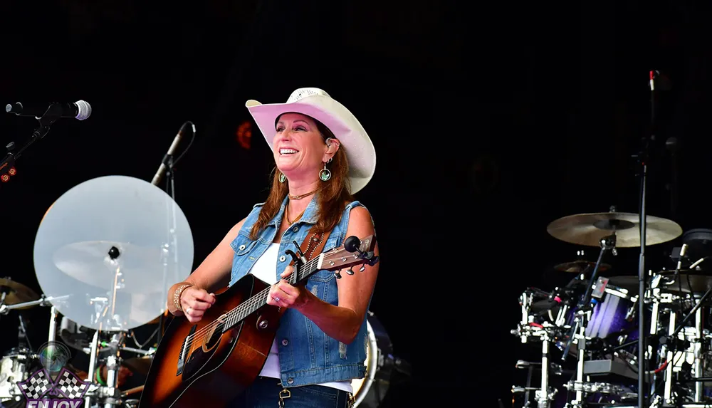 A smiling woman wearing a cowboy hat and denim vest is playing a guitar on stage with a drum set visible in the background