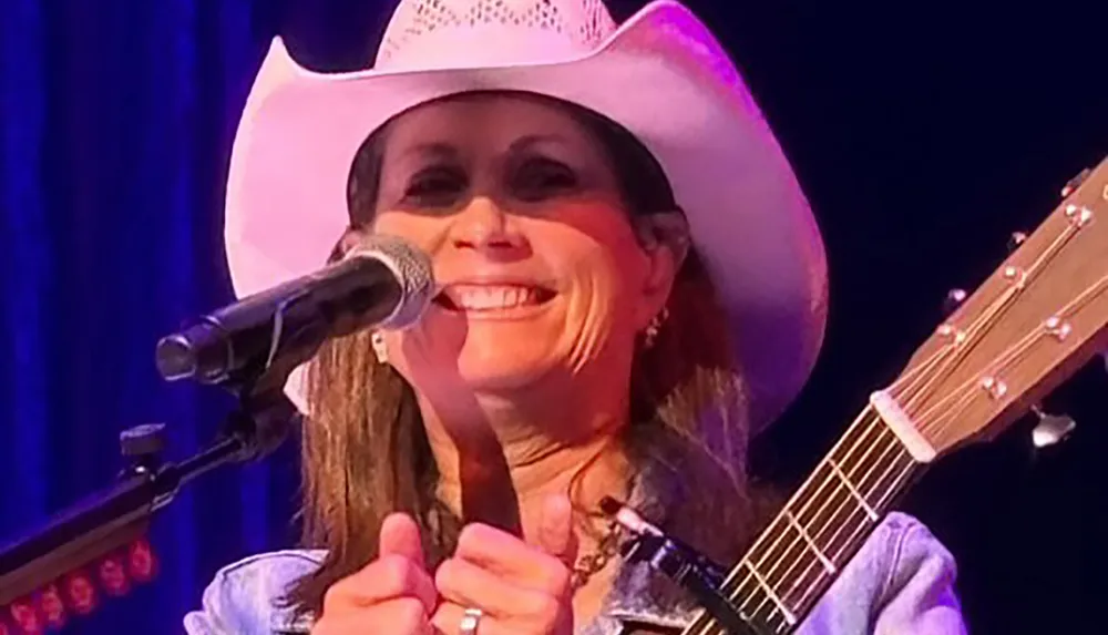 A person wearing a cowboy hat is smiling and standing with a microphone and guitar on stage