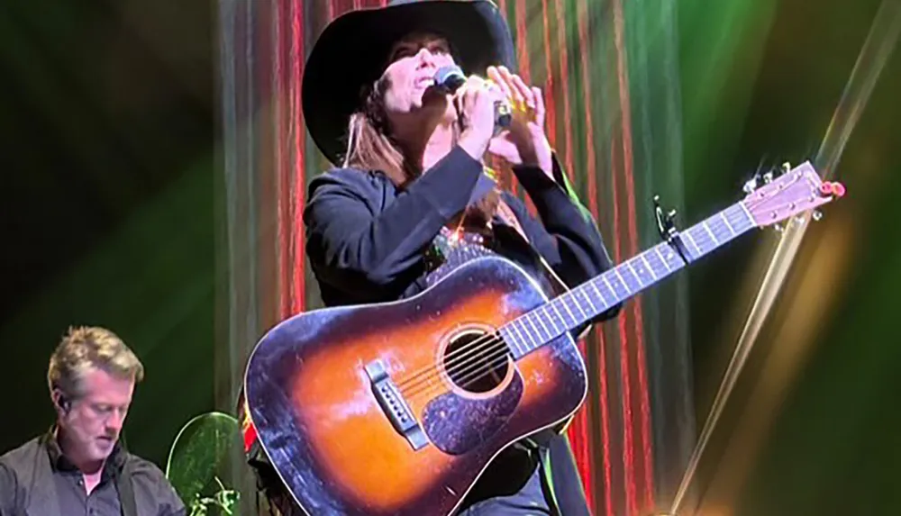 A musician in a black hat is playing a guitar and singing into a microphone on stage with dramatic lighting and a band member in the background