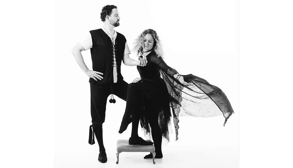 A man and woman pose dramatically in historical costumes with the woman standing on a small pedestal extending her dress