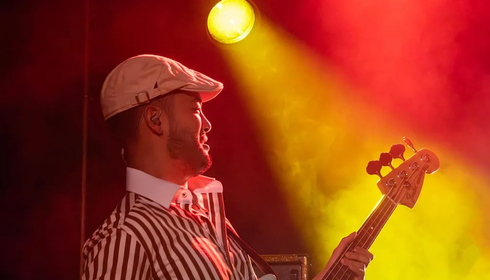 A musician wearing a striped shirt and cap plays a guitar under dramatic yellow and red stage lighting