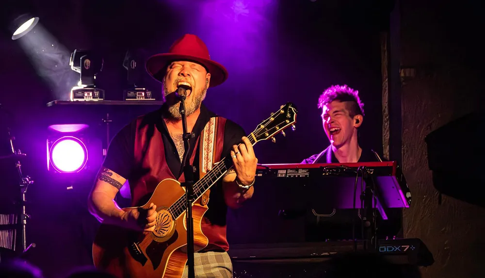 A musician passionately sings and plays guitar on stage beside a smiling keyboardist illuminated by vibrant purple lighting