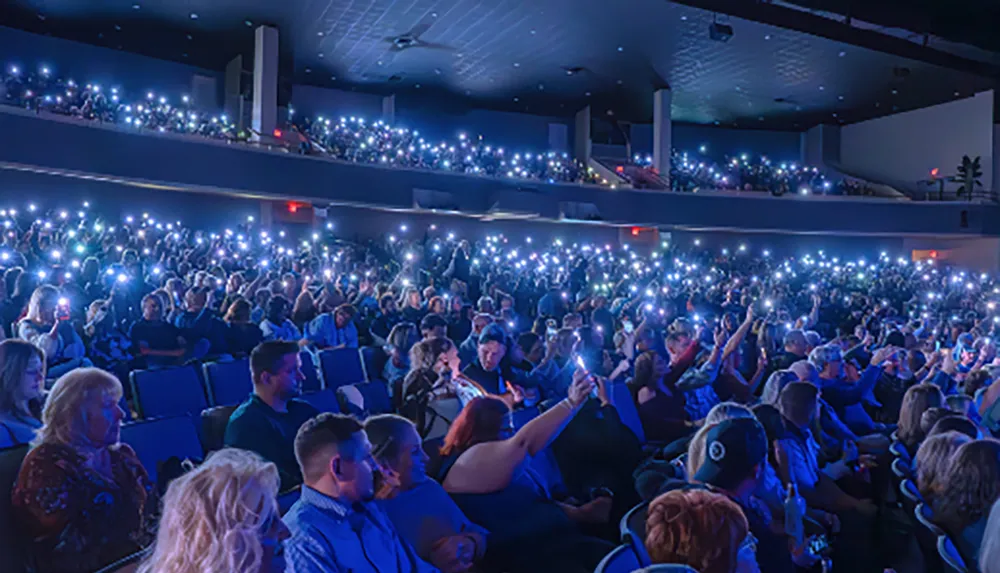 A large audience in a theater is holding up illuminated phones creating a sparkling effect in the dark