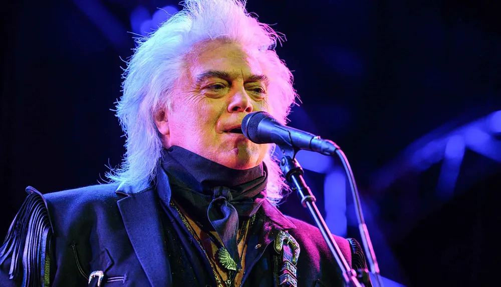 A performer with white hair is singing into a microphone under colorful stage lighting