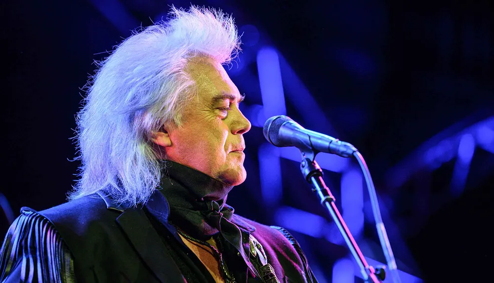 A person with silver hair is singing into a microphone on stage under vibrant blue lighting