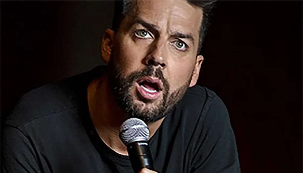 A man with a beard and short hair is speaking into a microphone with an animated expression