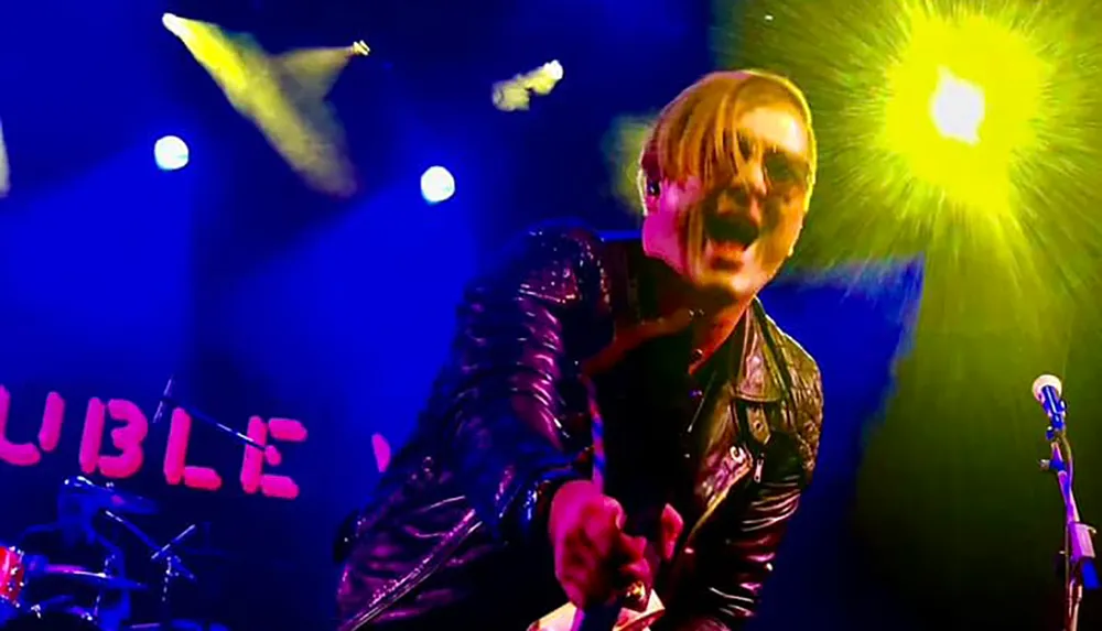 A person wearing sunglasses and a leather jacket energetically performs on stage under blue and yellow lights