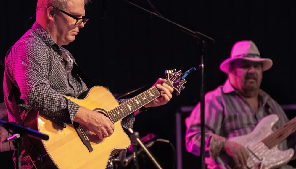 Two musicians are performing on stage one playing an acoustic guitar and the other playing a bass guitar with stage lighting highlighting them