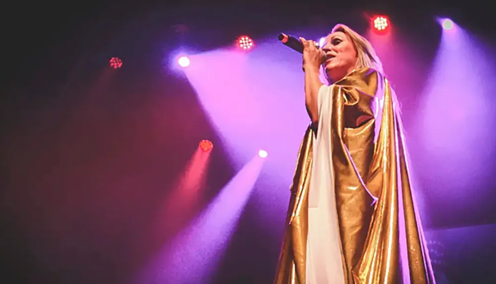 A person wearing a gold and white outfit is performing on stage under colorful lights