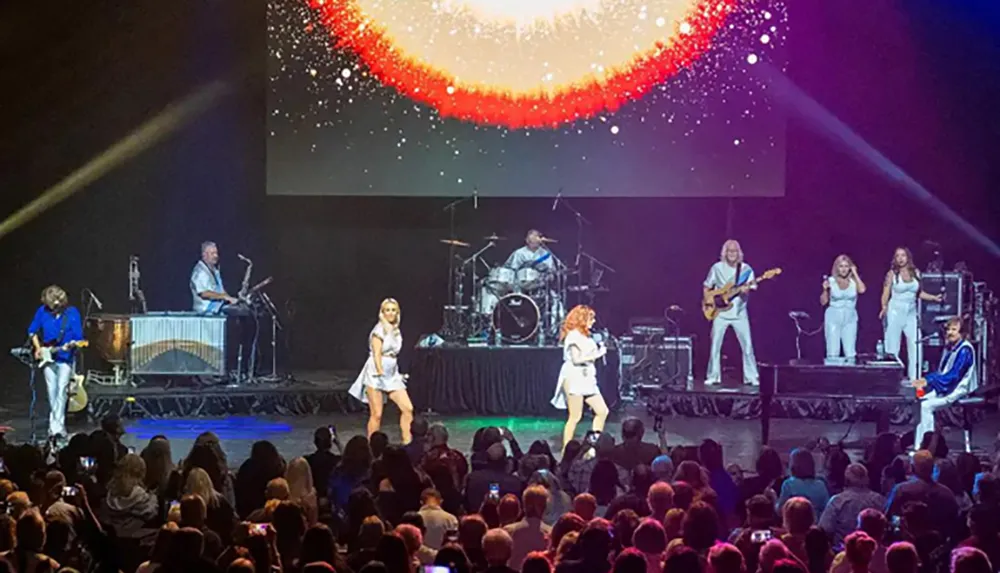 A band is performing energetically on stage in front of a large audience with a vibrant starry backdrop