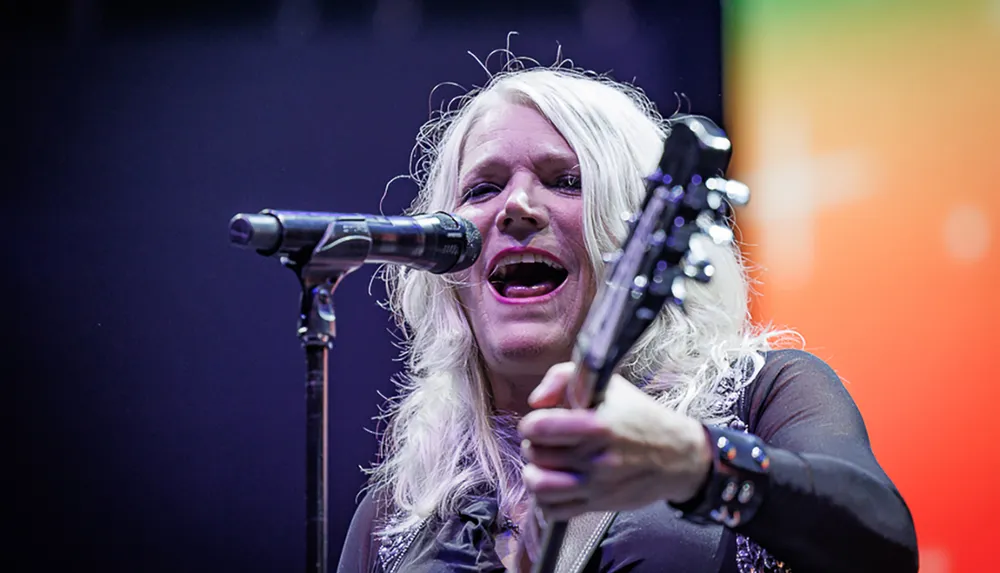 A woman with long hair is passionately singing into a microphone while playing a guitar on stage