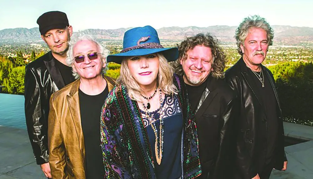 A group of five people dressed stylishly in jackets and hats pose together outdoors with a scenic landscape in the background