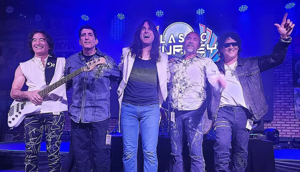 A group of five musicians stand together on stage in front of a Classic Journey sign smiling and posing for the camera