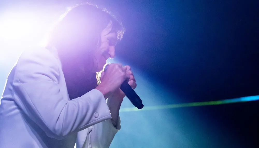 A person wearing a white jacket is passionately singing into a microphone on stage illuminated by bright lights