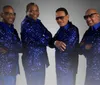 The image shows a group of seven men dressed in matching formal attire with four in white suits and three in blue sequined outfits posing against a dark background