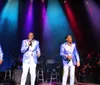 The image shows a group of seven men dressed in matching formal attire with four in white suits and three in blue sequined outfits posing against a dark background