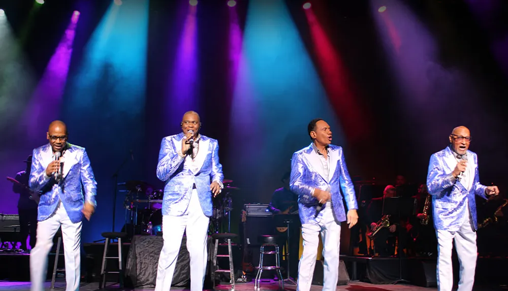 Four men in matching blue suits are performing on stage with colorful spotlights shining on them