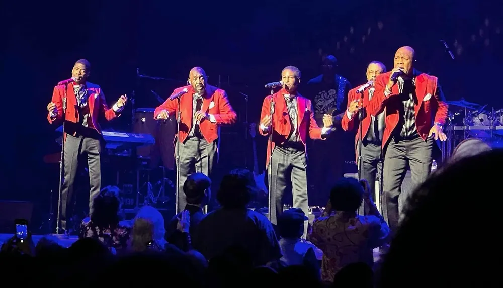 Five performers in matching red jackets sing on stage in front of an audience