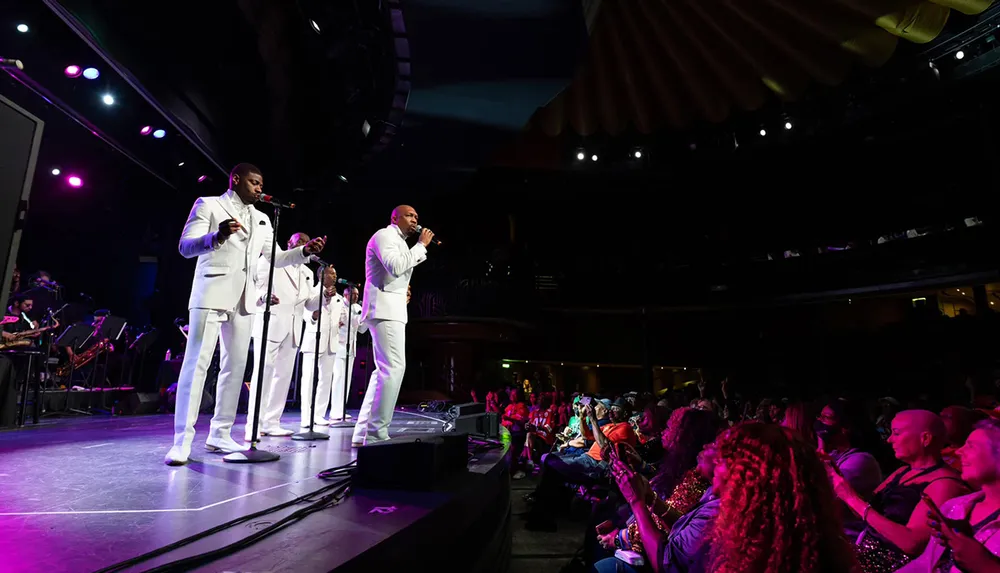 A group of singers in white suits perform on stage for an engaged audience in a dimly lit concert venue