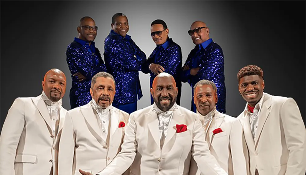 The image shows a group of seven men dressed in matching formal attire with four in white suits and three in blue sequined outfits posing against a dark background