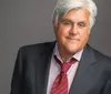 A man with gray hair is wearing a suit and red tie smiling at the camera against a gray background