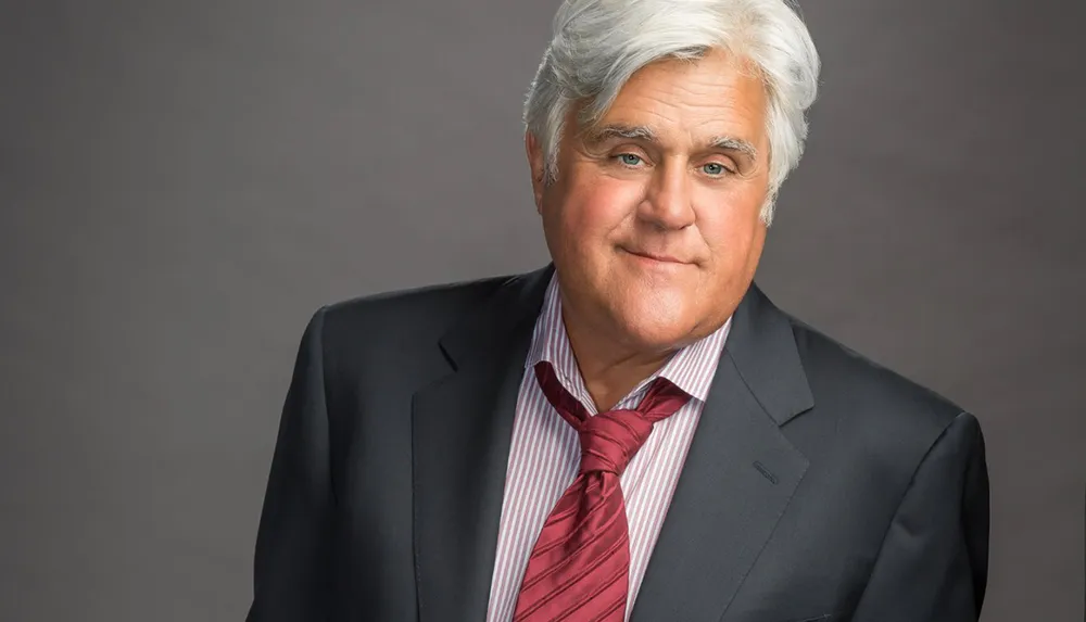 A man with gray hair is wearing a suit and red tie smiling at the camera against a gray background