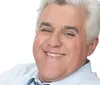 A man with gray hair is wearing a suit and red tie smiling at the camera against a gray background