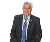 A man with gray hair is wearing a suit and red tie smiling at the camera against a gray background