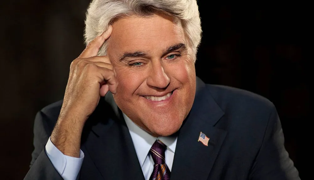 A smiling man with gray hair in a suit is posing with his hand on his temple