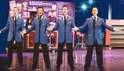 Four performers in matching blue suits are singing on stage in front of a bar-themed backdrop.