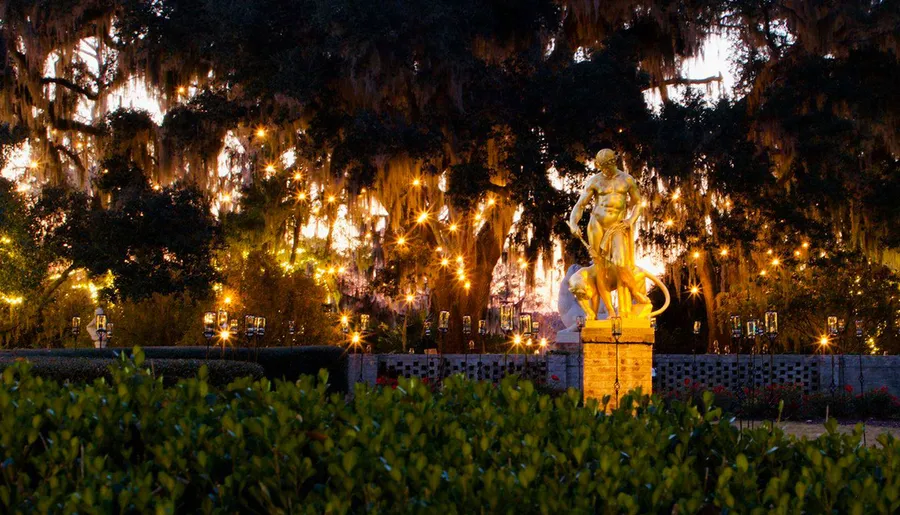 The image shows a gilded statue bathed in warm light, set against a backdrop of lush trees draped with Spanish moss and illuminated by twinkling lights as evening falls.