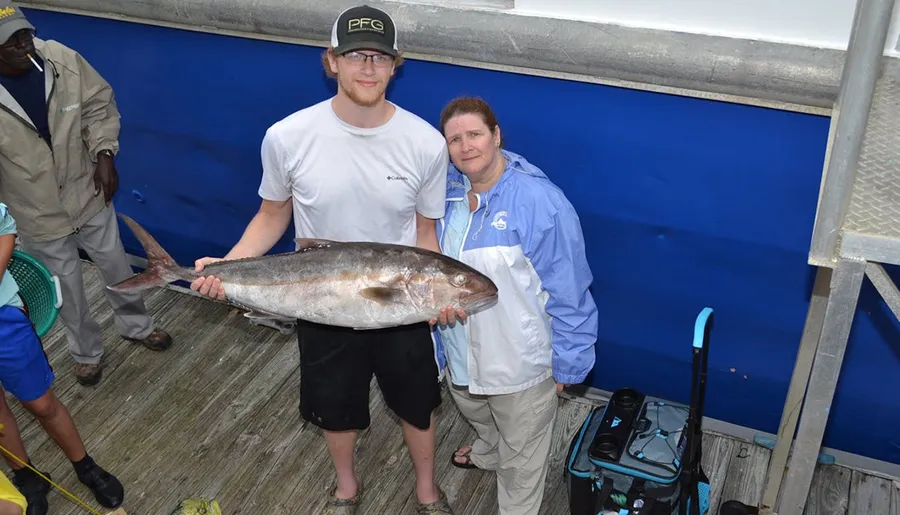 Two people are posing with a large fish they have caught, standing on a wooden pier with a blue wall in the background.