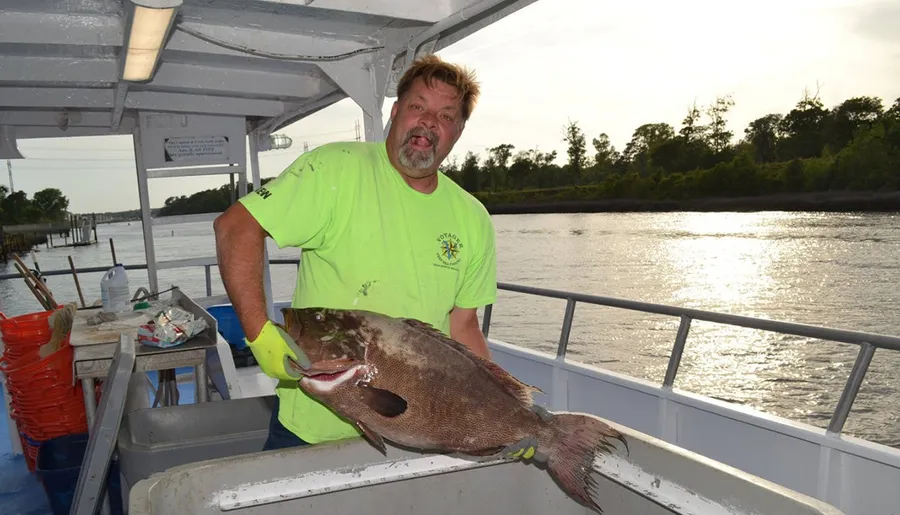 A man in a neon green shirt is playfully sticking out his tongue while holding a large fish on a boat with a river and trees in the background.