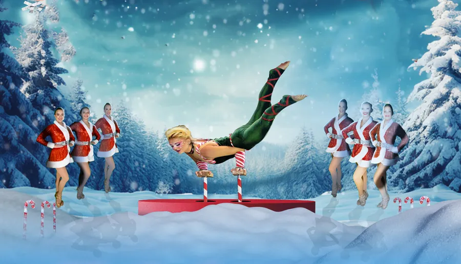 A performer in festive attire balances on candy cane stands surrounded by dancers dressed in Santa costumes against a snowy forest backdrop.