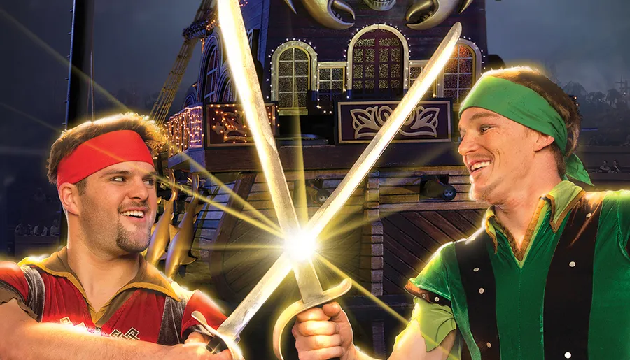 Two people dressed as fantasy characters are playfully crossing swords in front of a whimsical backdrop, resembling a scene out of a theatrical or theme park setting.