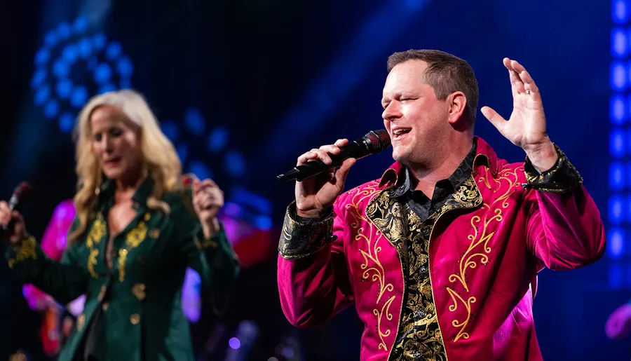 Two singers wearing vibrant traditional jackets are performing on stage with one grasping a microphone and gesturing expressively.