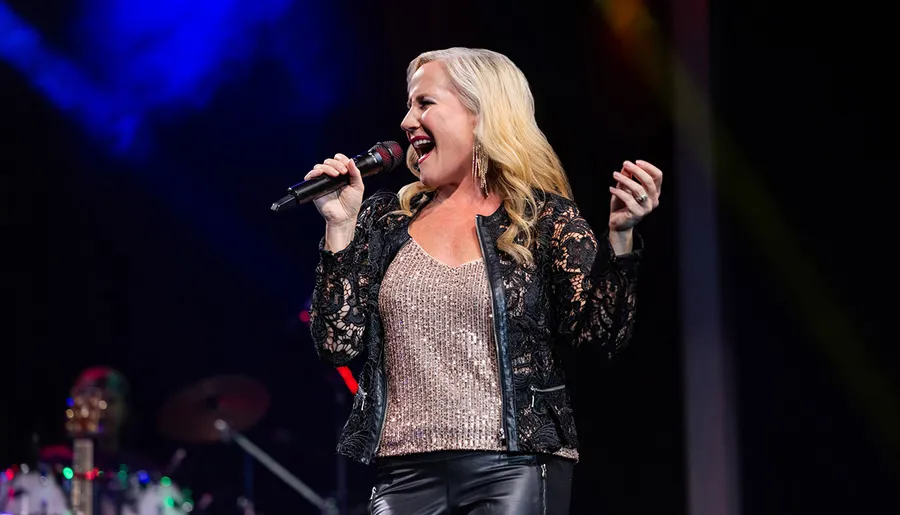 A female singer is performing on stage, holding a microphone and singing with passion under colorful stage lighting.