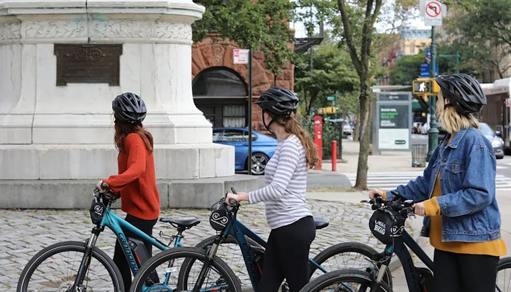 Three people wearing helmets stand with bicycles near a stone monument in an urban area