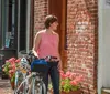 A woman wearing a pink top stands with her bicycle on a brick sidewalk next to flower pots as a man walks by in the background