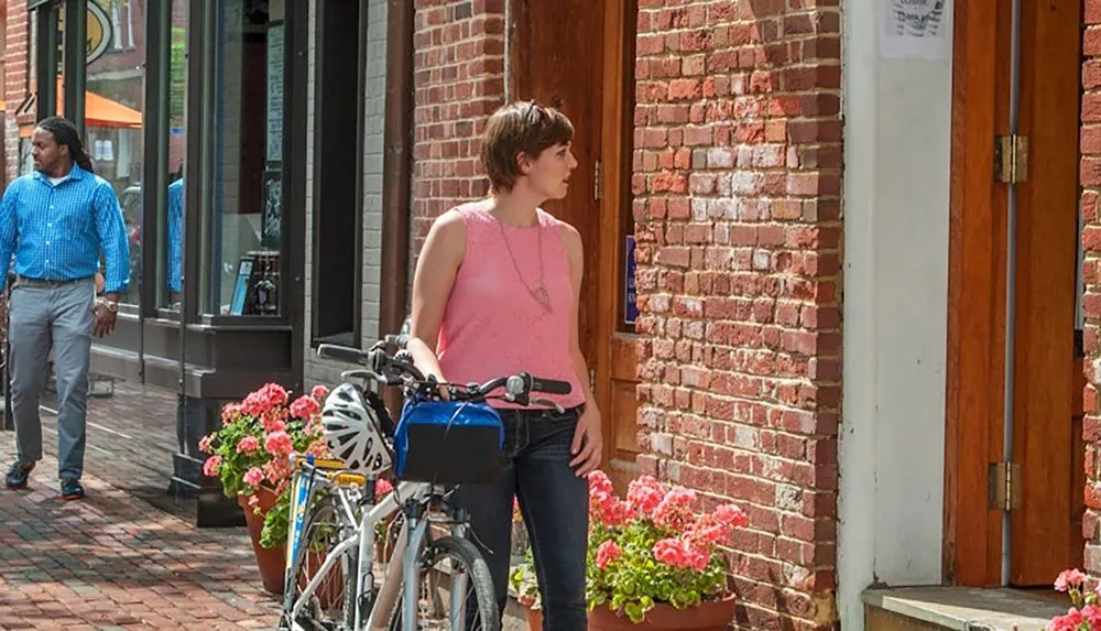 A woman wearing a pink top stands with her bicycle on a brick sidewalk next to flower pots as a man walks by in the background