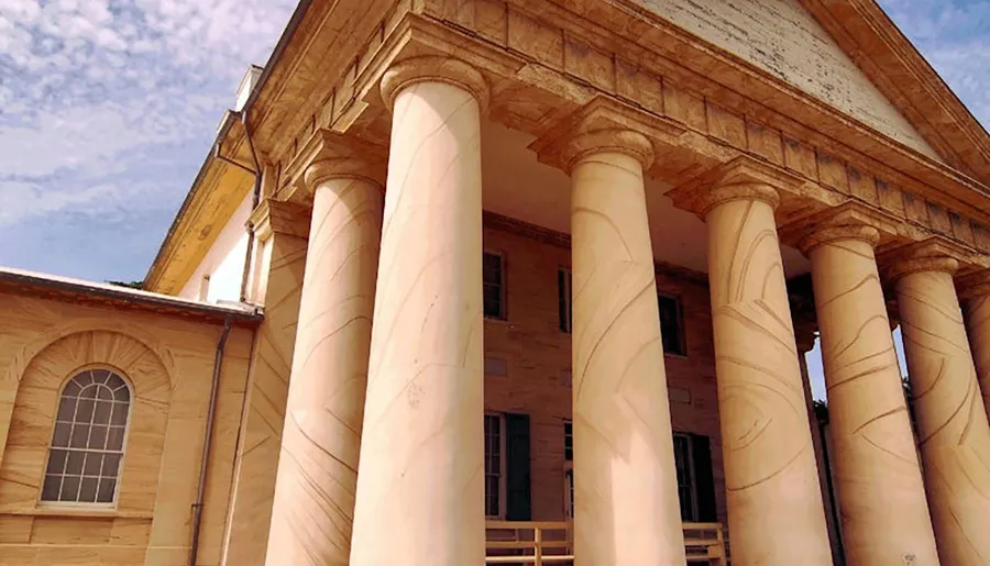 The image shows a classical building with large stone columns and a triangular pediment against a partly cloudy sky.