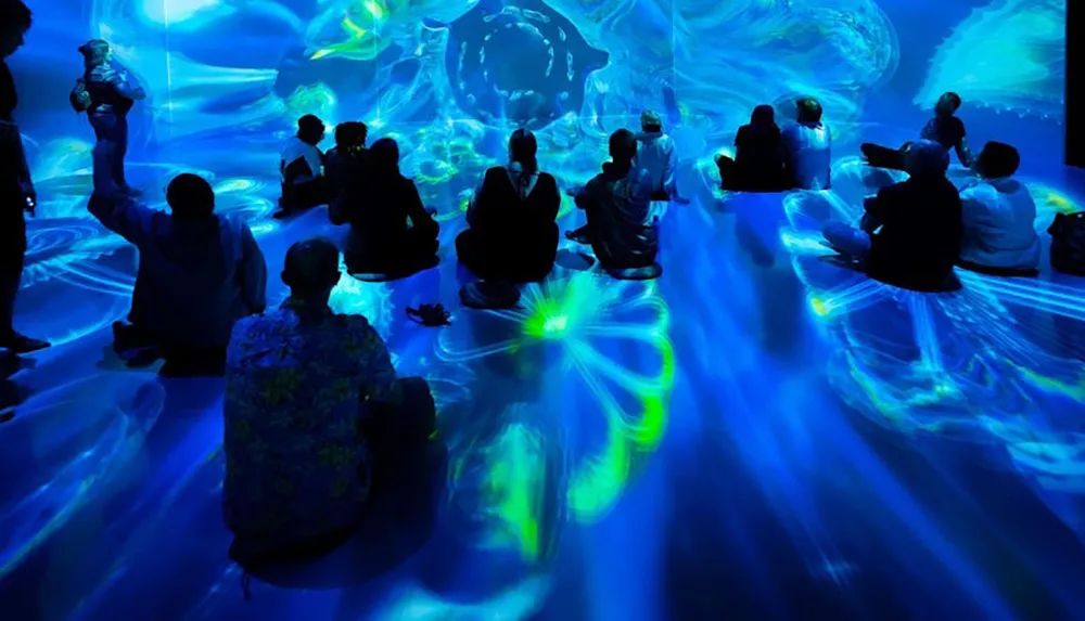 A group of people is seated in a room illuminated with blue and green abstract light projections