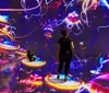 Two people stand and sit on platforms amidst a vibrant immersive digital art installation with colorful abstract jellyfish-like forms