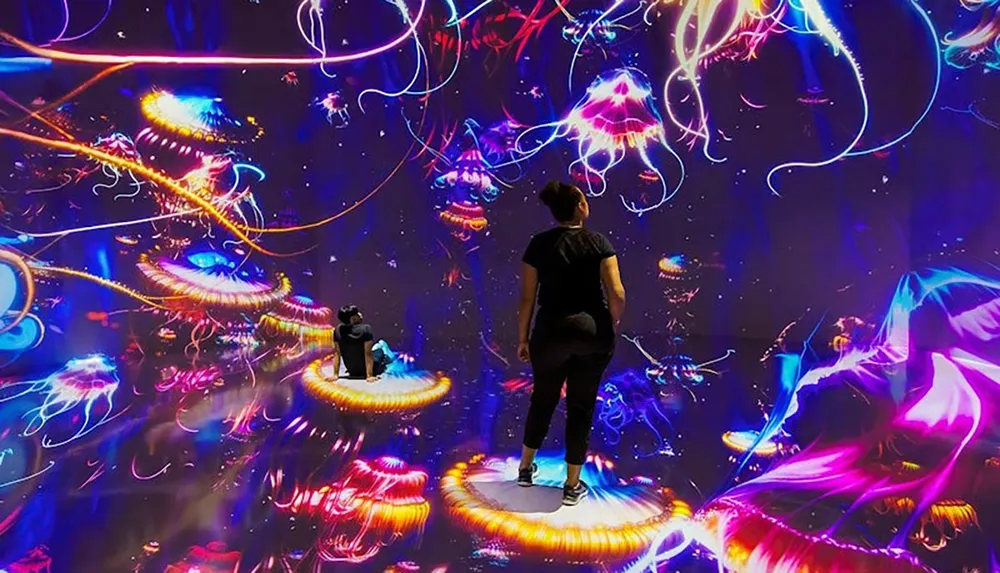 Two people stand and sit on platforms amidst a vibrant immersive digital art installation with colorful abstract jellyfish-like forms