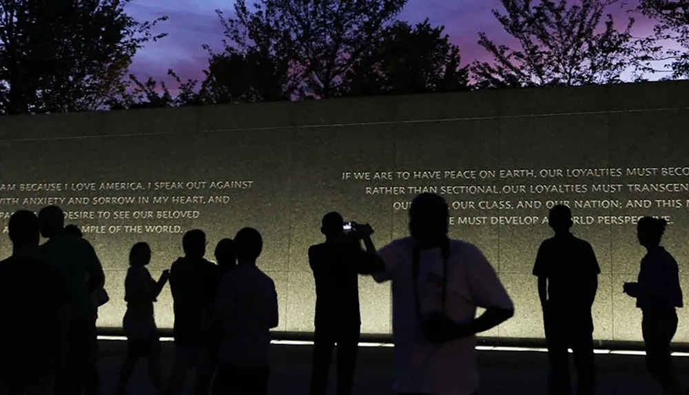 People are silhouetted against a wall with engraved text possibly at a memorial or monument during twilight