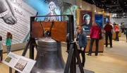 Visitors are exploring a museum exhibit featuring the Liberty Bell with historical displays in the background.