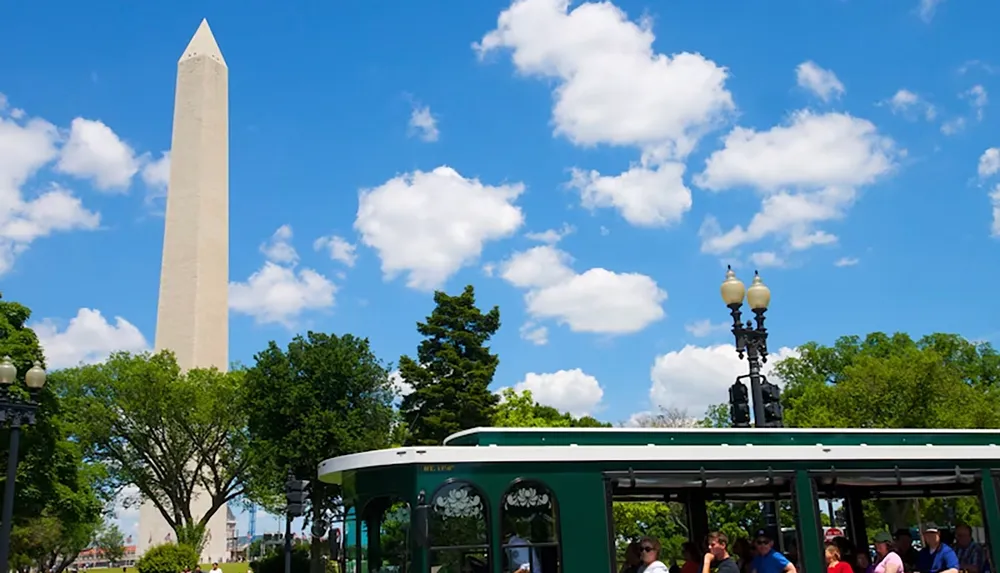 The image shows the Washington Monument against a backdrop of a bright blue sky with fluffy clouds and people riding a green trolley in the foreground