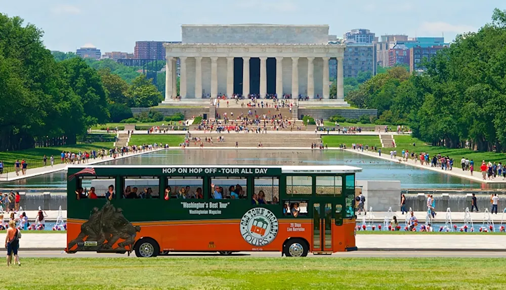 A tour trolley passes by the Lincoln Memorial with people walking around the reflecting pool in Washington DC